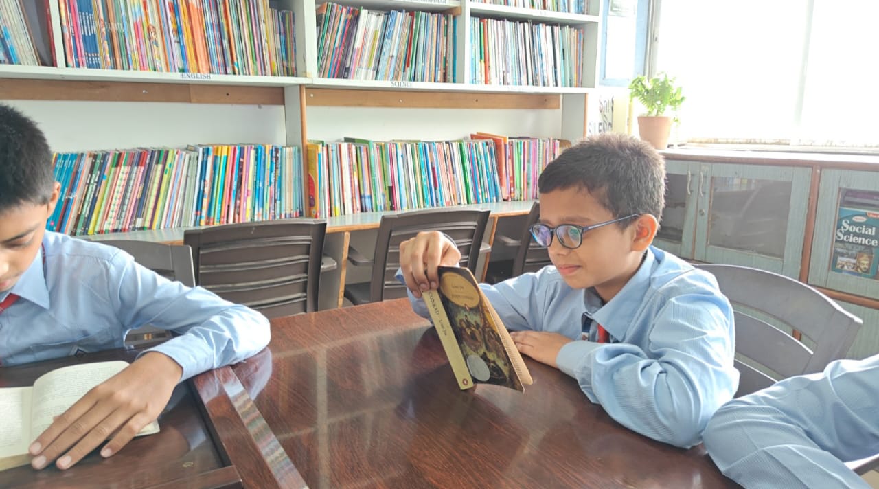 A student wearing glasses reading a book in the library of a Top CBSE school in Jayanagar. Bookshelves filled with colorful books are in the background.