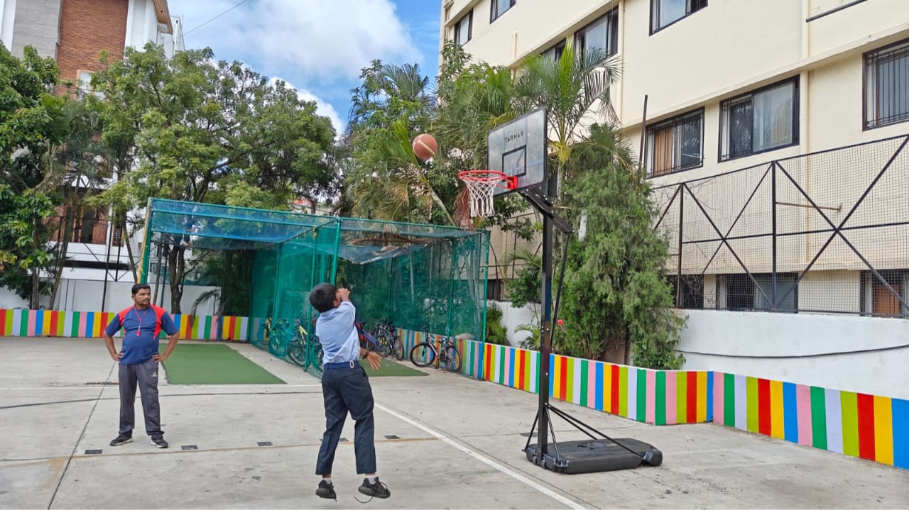 A student shooting a basketball at the outdoor court of Alpine School, a Popular CBSE school in Banashankari, with a coach observing nearby.